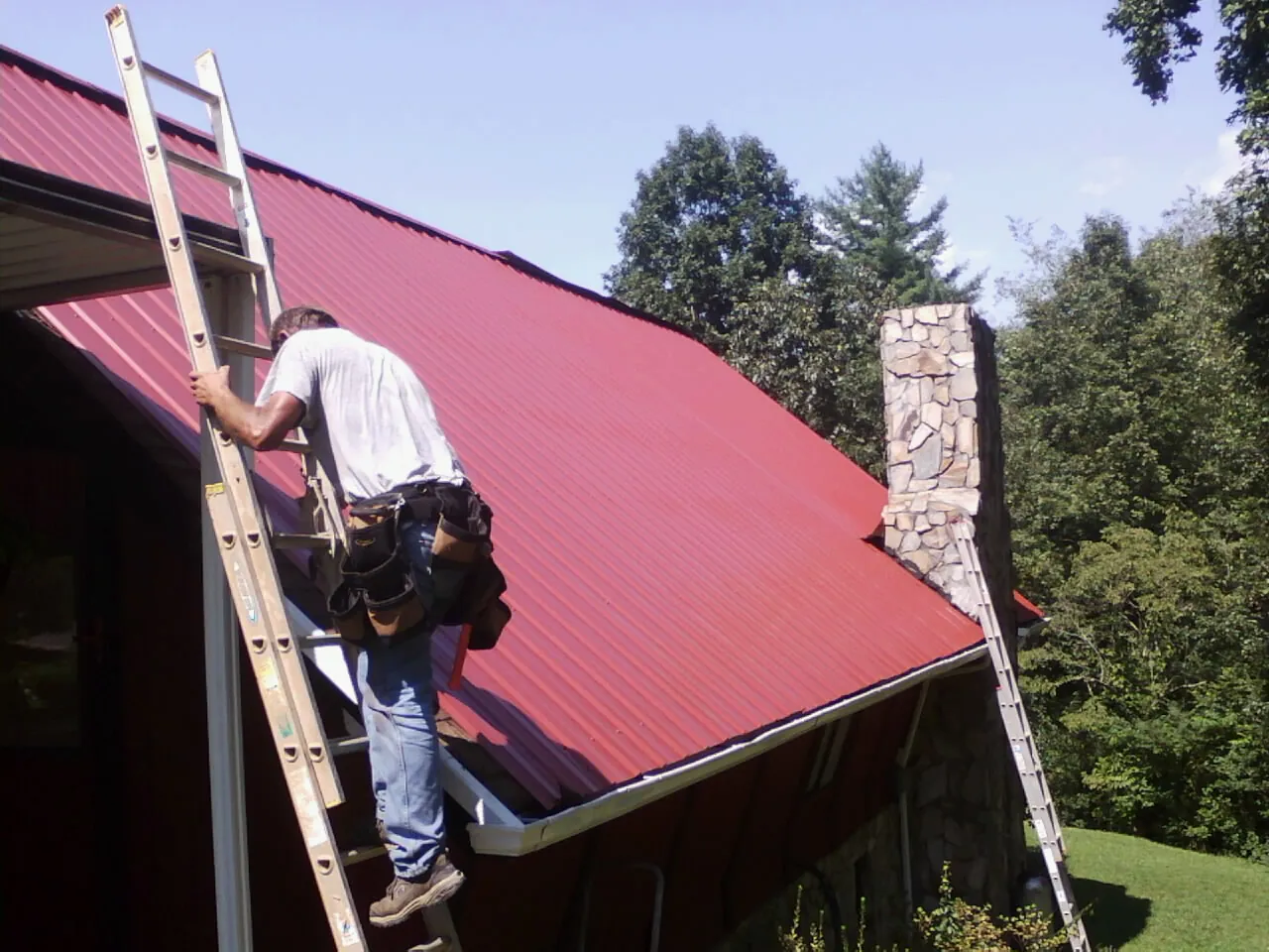 Roofer on a ladder beside a red metal roof and stone chimney — Soffit & Fascia Repair by Roofing Brothers in Lafferty